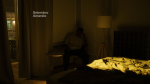 A imagem mostra um homem sentado ao lado da cama com a mão na cabeça, é noite lá fora