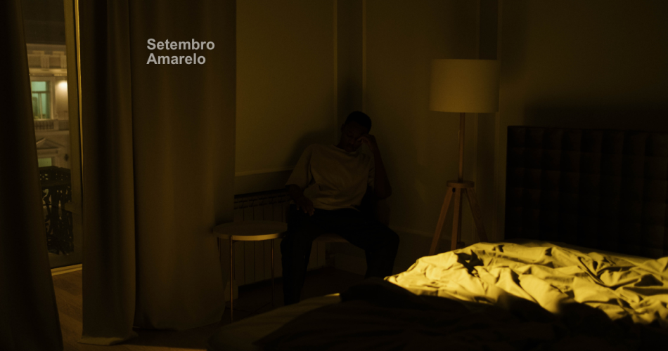 A imagem mostra um homem sentado ao lado da cama com a mão na cabeça, é noite lá fora
